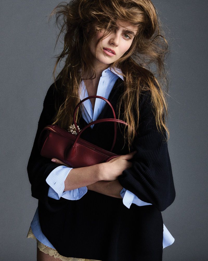 Adult female fashion model poses in a photo studio with a red leather purse, wearing a blue button-down shirt and black sweater.