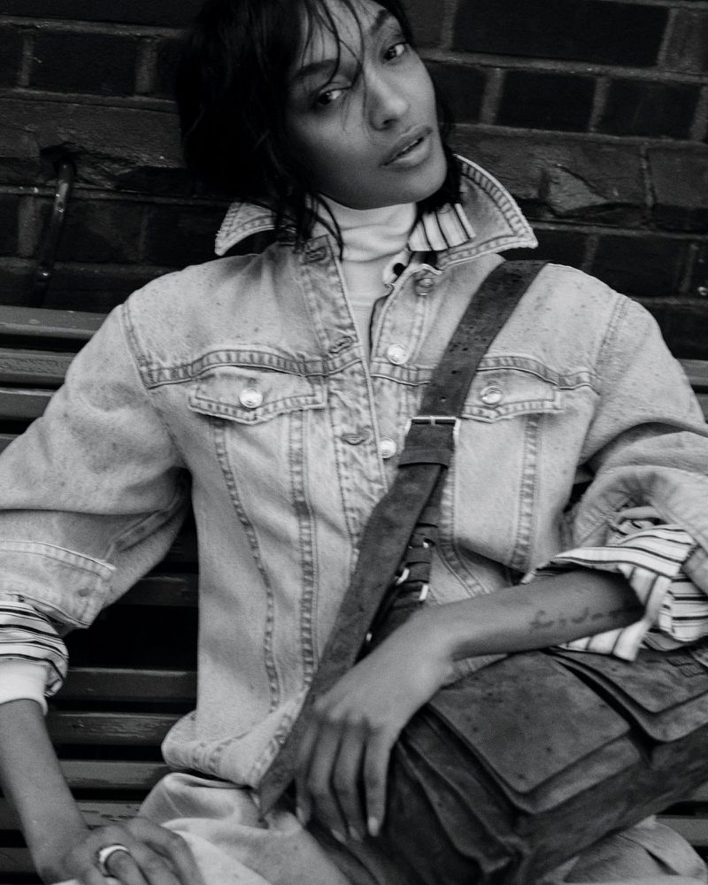 A woman with dark hair wearing a denim jacket, turtleneck, and carrying a satchel, poses seated against a brick wall backdrop.
