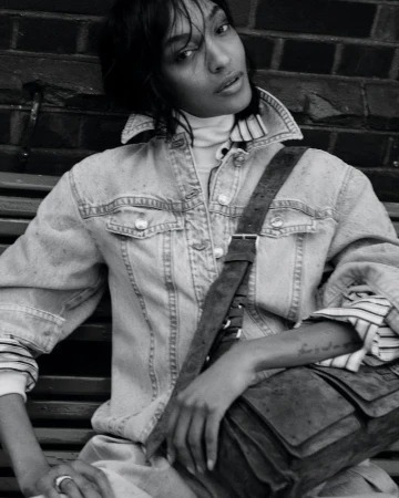 A woman with dark hair wearing a denim jacket, turtleneck, and carrying a satchel, poses seated against a brick wall backdrop.