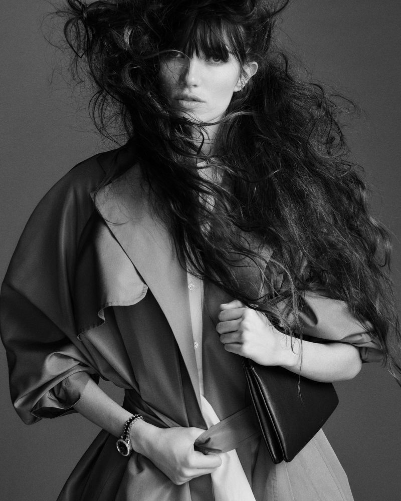 Black and white portrait of a woman with long hair and a trench coat, holding a clutch bag in a studio setting with a neutral background.