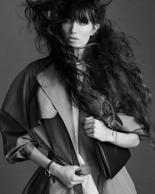 Black and white portrait of a woman with long hair and a trench coat, holding a clutch bag in a studio setting with a neutral background.