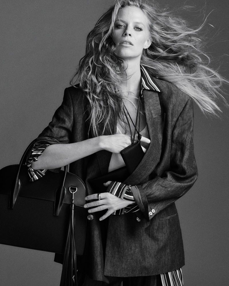 Model with long blowing hair wearing an oversized blazer and holding a black handbag against grey seamless background.