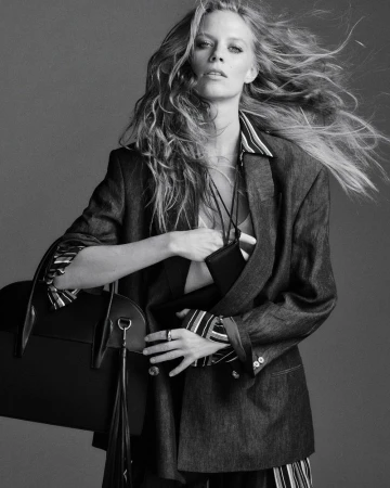 Model with long blowing hair wearing an oversized blazer and holding a black handbag against grey seamless background.