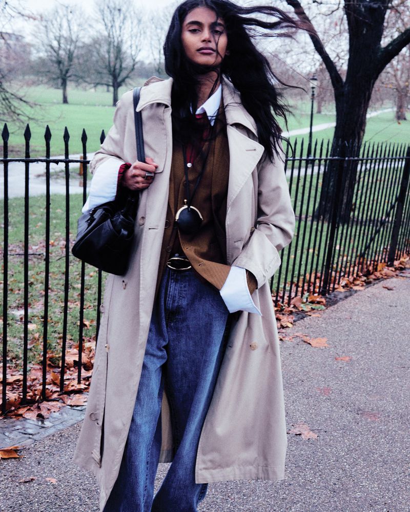 A woman in a trench coat walks in a park on a cloudy day. She carries a black purse over her shoulder and wears jeans and a brown jacket.
