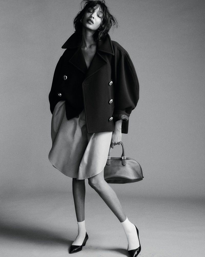 Model in dark double-breasted coat holding handbag against a seamless grey studio background