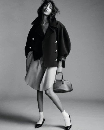 Model in dark double-breasted coat holding handbag against a seamless grey studio background