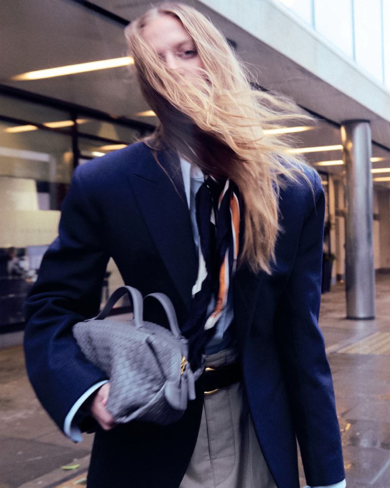 Fashion portrait featuring an adult woman with long blond hair carrying a grey handbag, styled in a navy blazer and scarf outdoors.