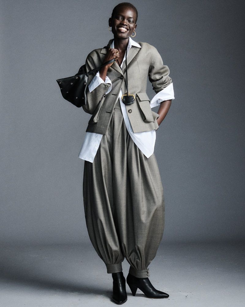 Fashionable African woman posing in a grey suit and black heeled boots holding a studded leather bag in a grey studio environment.