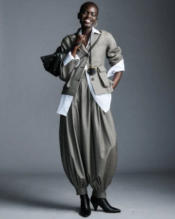 Fashionable African woman posing in a grey suit and black heeled boots holding a studded leather bag in a grey studio environment.