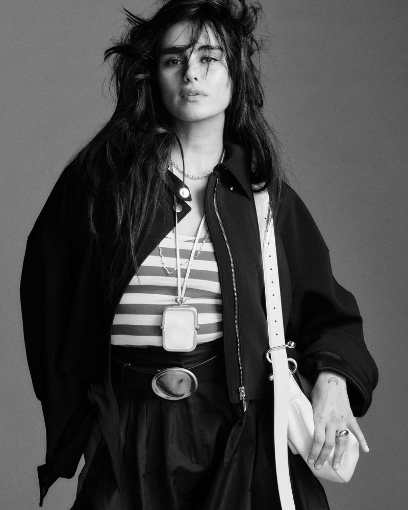 Model in dark jacket and striped top wearing a shoulder bag against a grey studio backdrop for British Vogue.