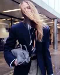 Model in a navy blazer holding a grey handbag with long blonde hair blowing across her face outdoors