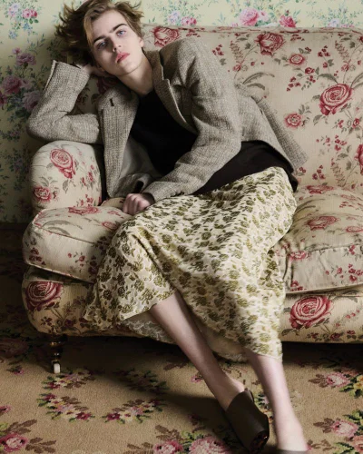 Model seated on a floral patterned sofa wearing a tweed blazer, black top, and floral skirt for British Vogue.