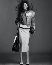 Model with wild dark hair wearing a patterned blazer and white skirt against a grey background
