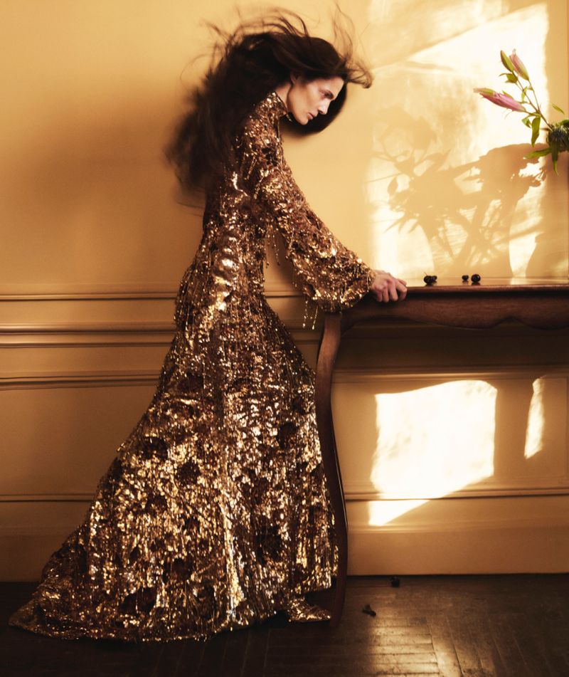 Woman in long gold dress stands in front of a table with a vase of flowers. She is leaning forward and looking down.