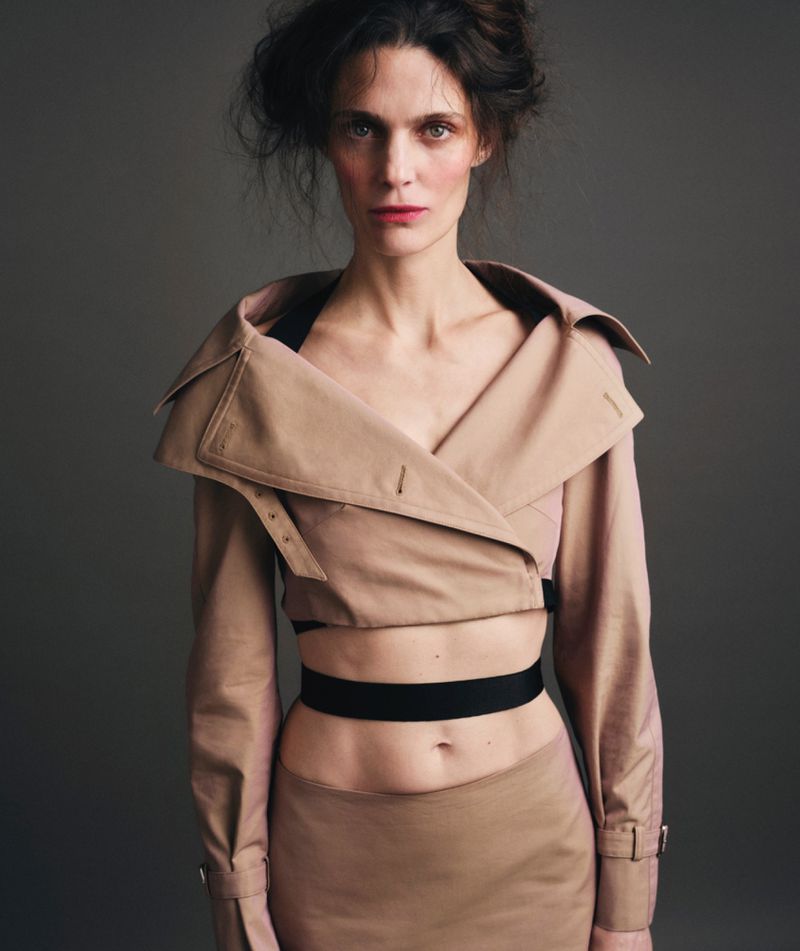 A woman in a beige crop top jacket and skirt looks directly at the camera against a neutral background in a fashion portrait.