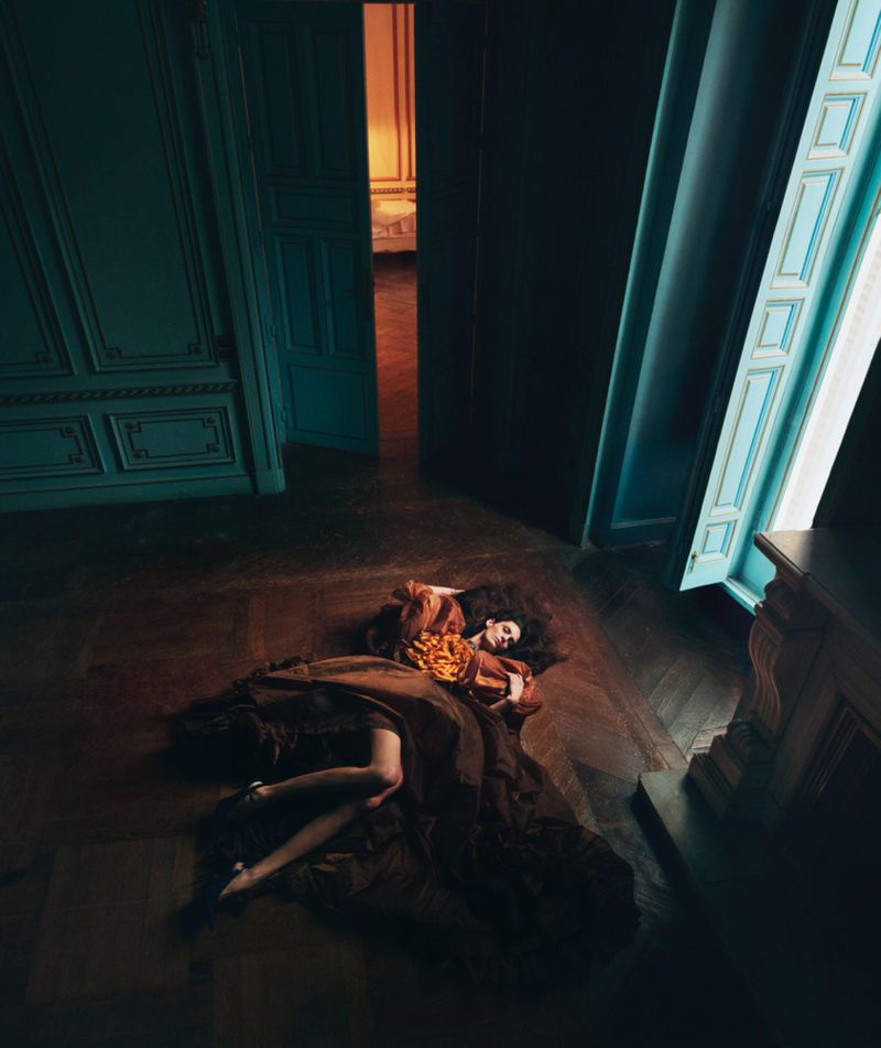 Adult woman in flowing brown gown lies on a wood floor in a room with paneled walls and open door to room with bed.