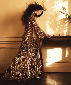 Model in a long sequined gold dress standing by a wooden table with flowers against a warm interior wall.
