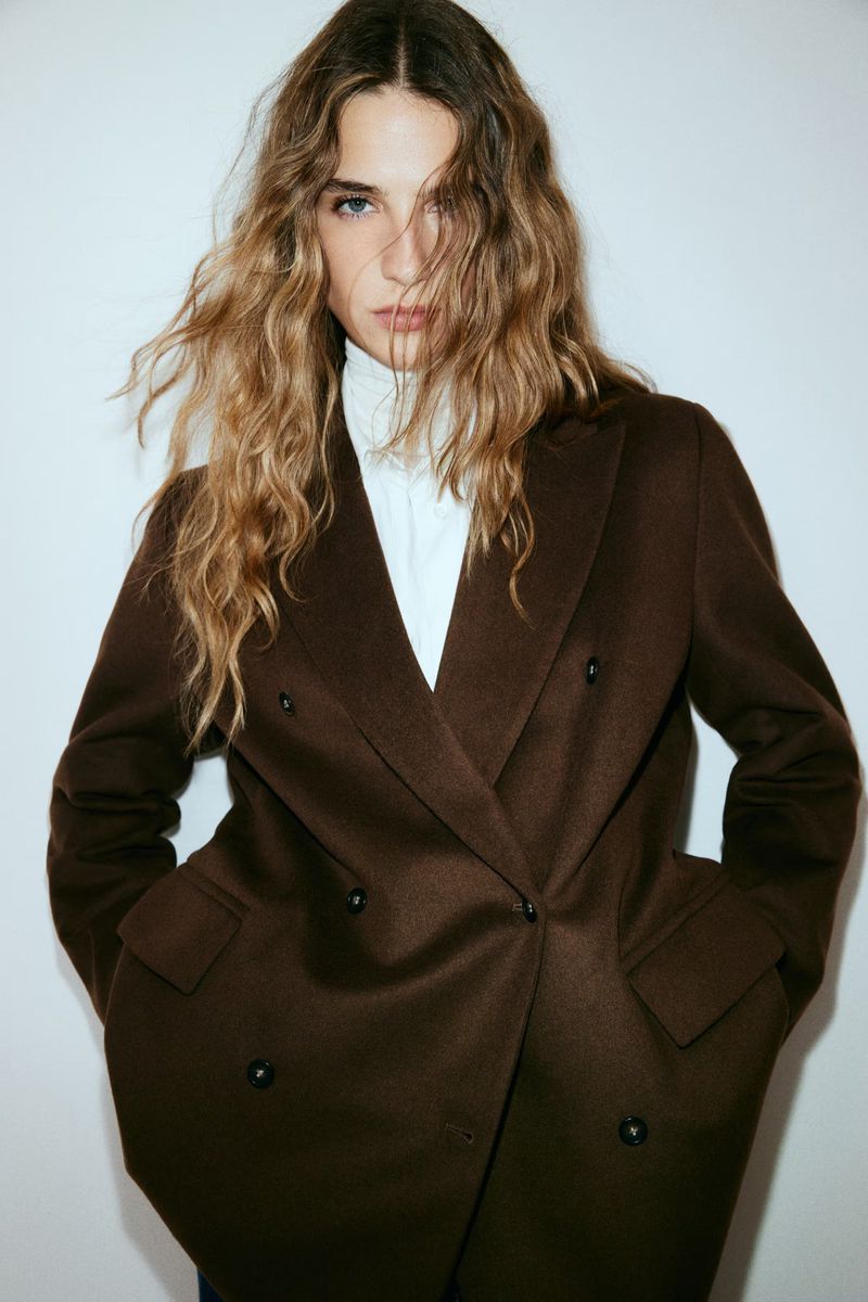 A woman with long, wavy blonde hair poses indoors wearing a brown, double-breasted blazer with a white top against a white backdrop.