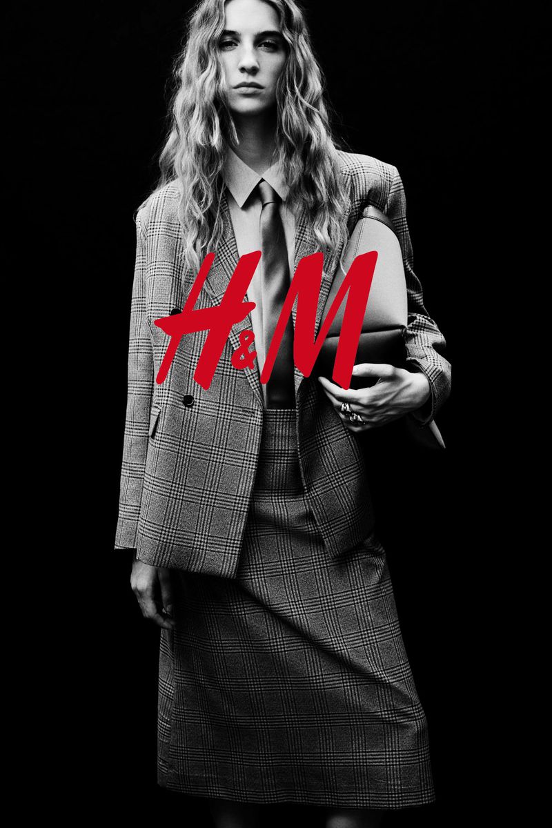 Fashion photo of woman with long blonde hair wearing plaid suit, collared shirt, and tie, holding a clutch against a black backdrop.