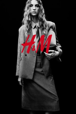 Fashion photo of woman with long blonde hair wearing plaid suit, collared shirt, and tie, holding a clutch against a black backdrop.