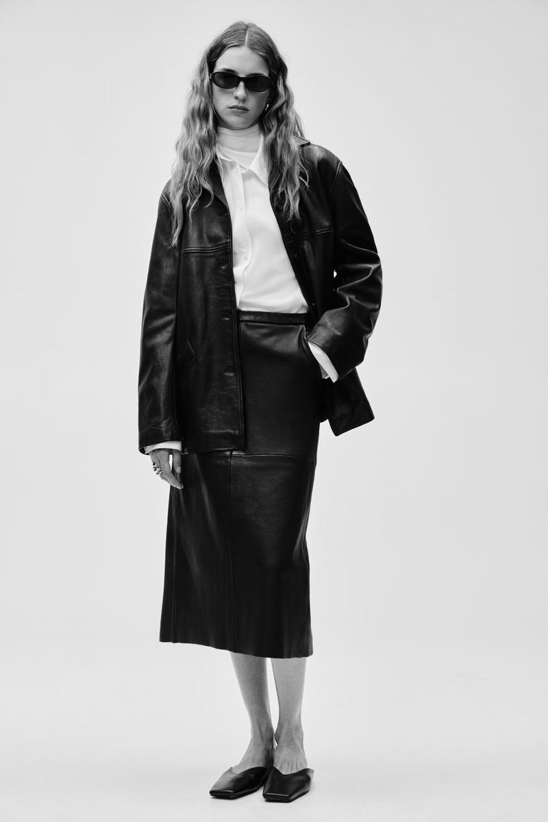 Black and white fashion shot of a woman wearing a black leather skirt suit, white turtleneck and blouse, and black flats on a white backdrop.