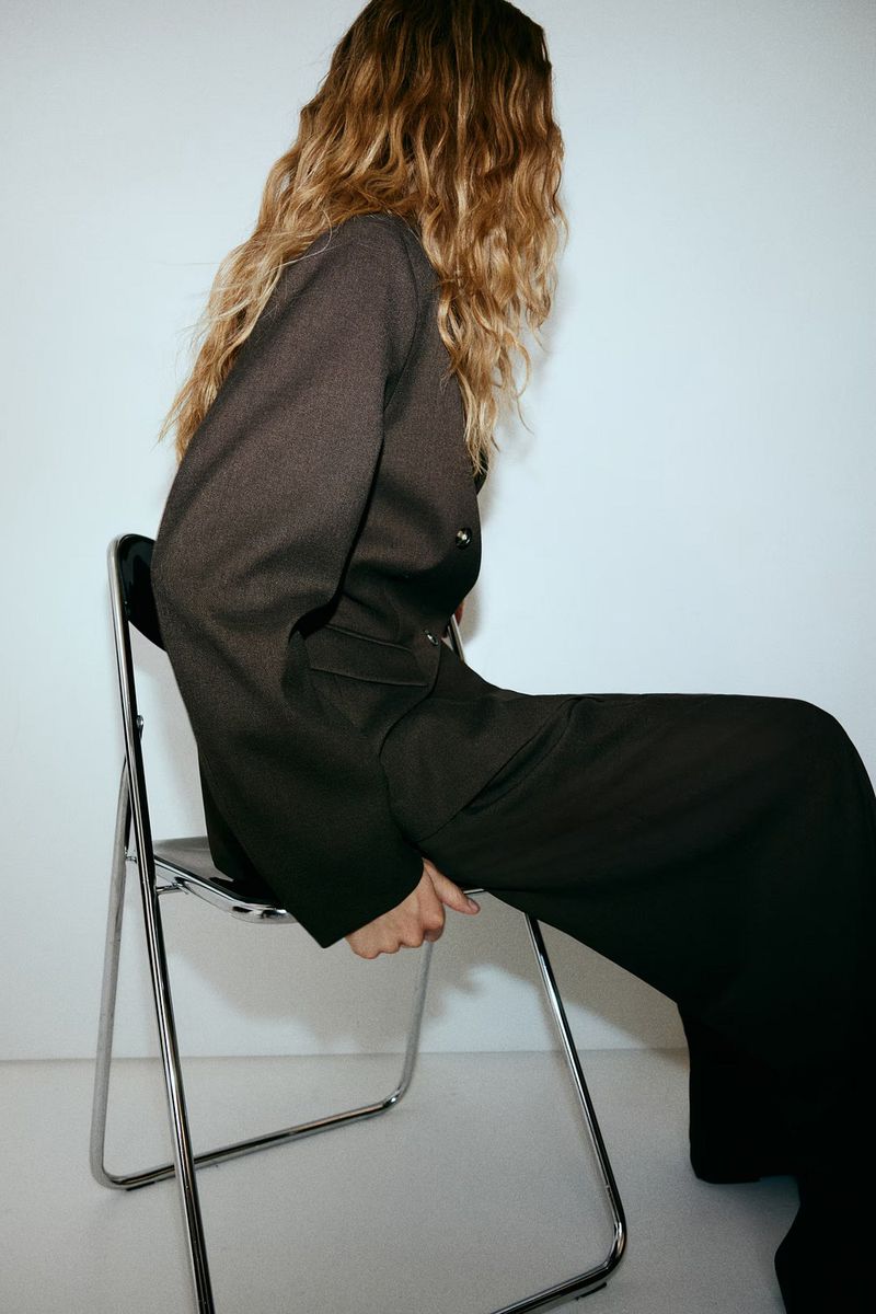 Blonde woman in a dark grey blazer and dark pants sits on a chrome folding chair in a light studio setting against a white wall.
