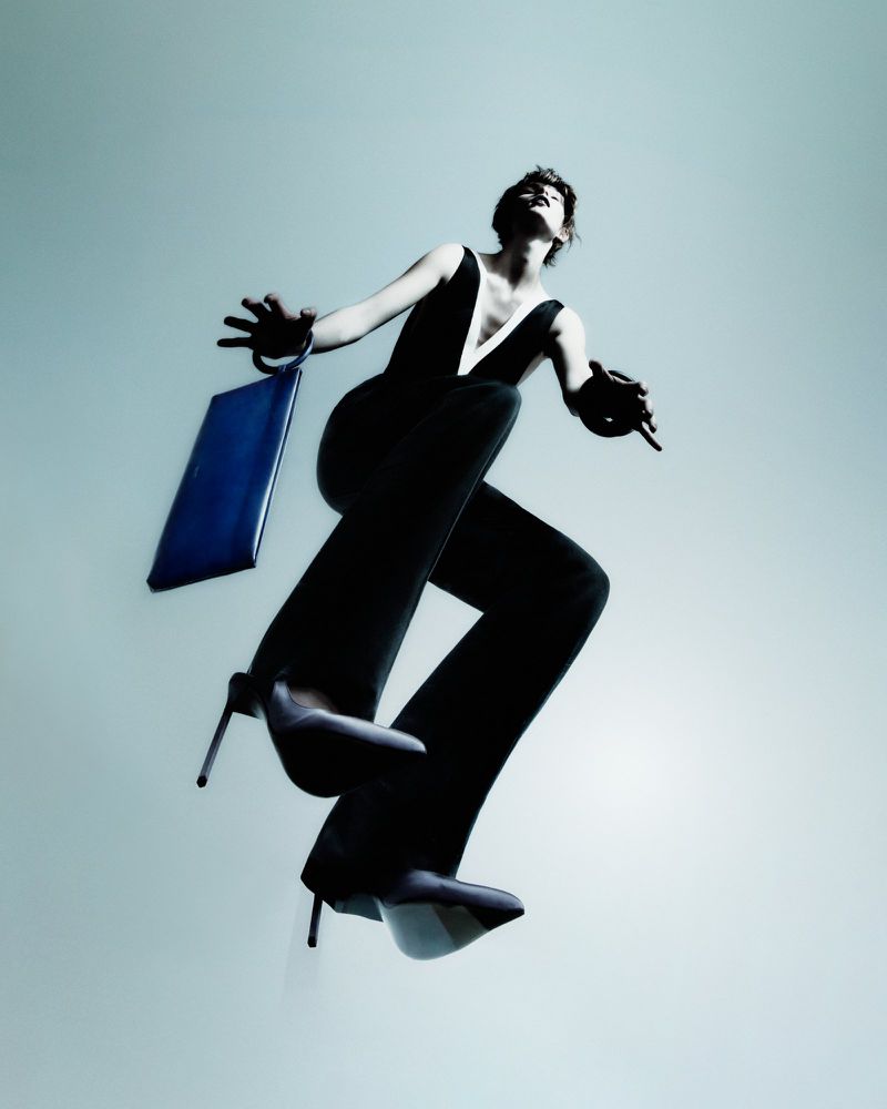 Model in black jumpsuit wearing high heels holding a blue handbag in mid air against a light grey studio background