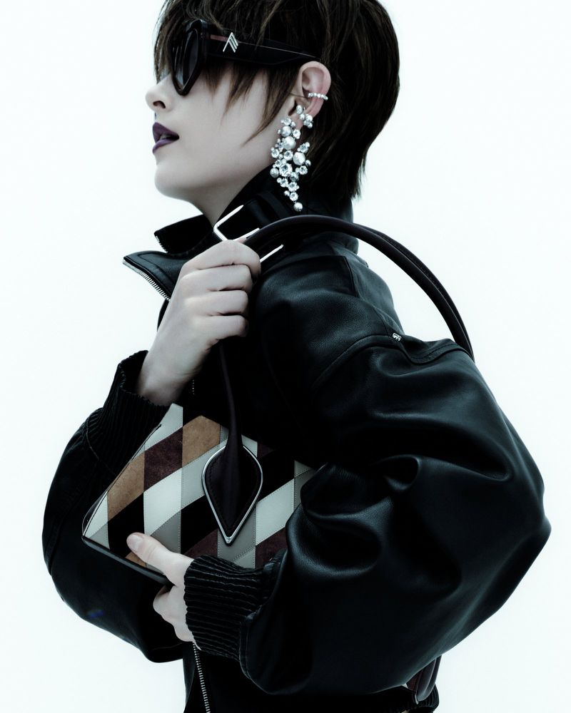 Profile portrait of a short-haired model wearing a black leather jacket and pearl earrings, holding handbags against a white background.