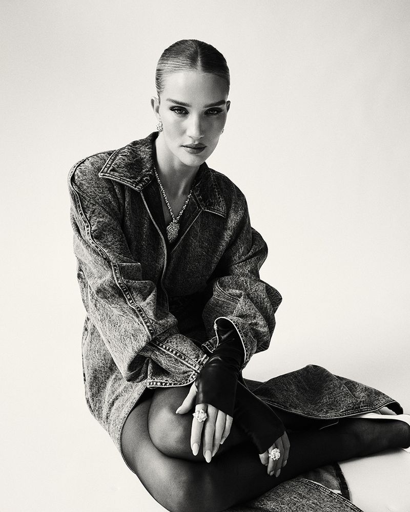 Monochrome studio portrait of a woman with slicked-back hair wearing a denim jacket, black gloves, and statement jewelry, seated on the floor.