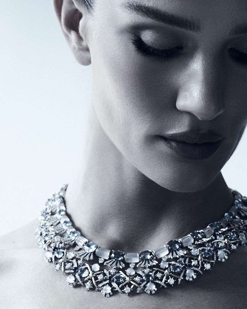 Close-up portrait of a woman with her eyes closed, wearing a large, elaborate jeweled necklace against a light blue gradient background.