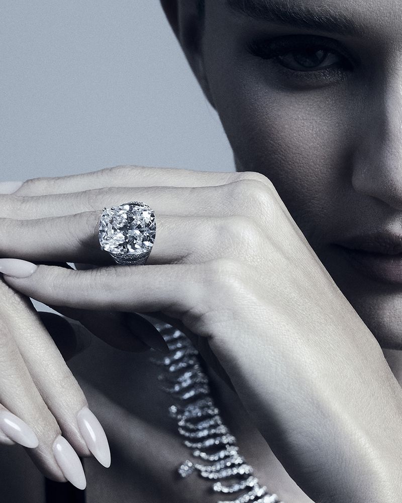 Close-up of an adult woman's face and hands, featuring a large diamond ring and a diamond necklace against a light gray backdrop.