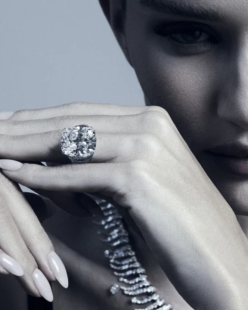 Close-up of an adult woman's face and hands, featuring a large diamond ring and a diamond necklace against a light gray backdrop.