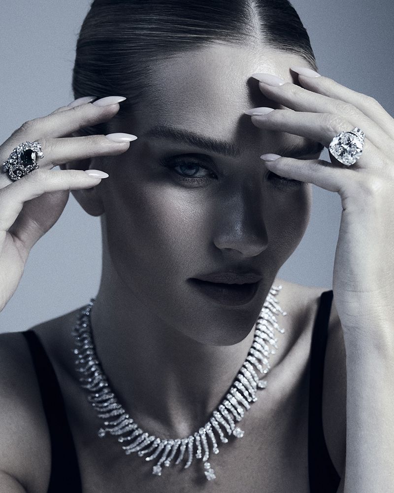 Close-up shot of an adult woman wearing an ornate diamond necklace and diamond rings, her hands near her face in a studio setting.
