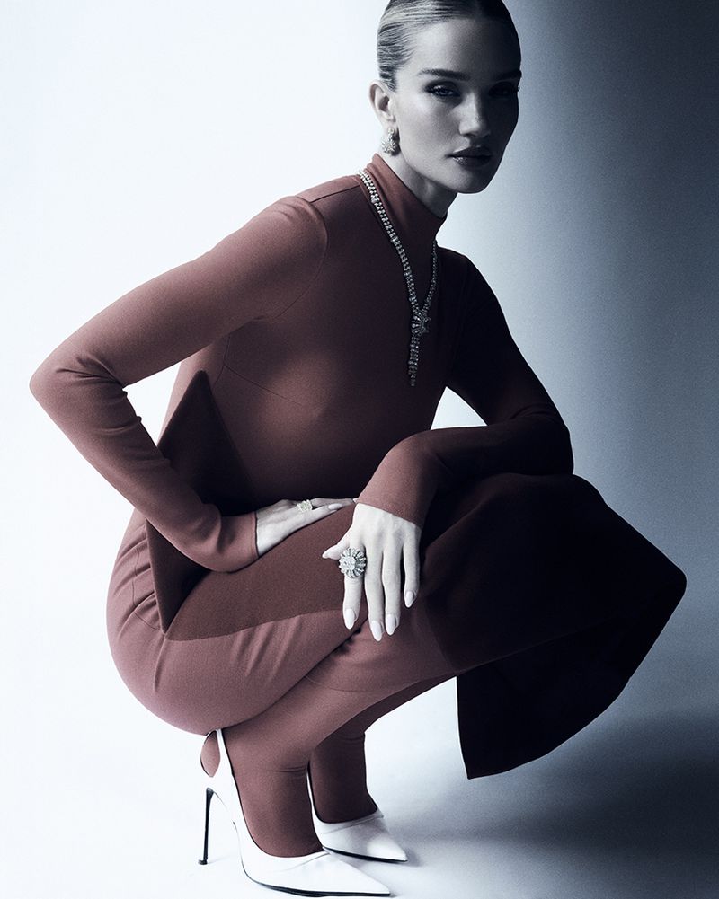 Elegant woman in a terracotta turtleneck jumpsuit poses while crouching, complemented by a white stiletto heel and statement jewelry.