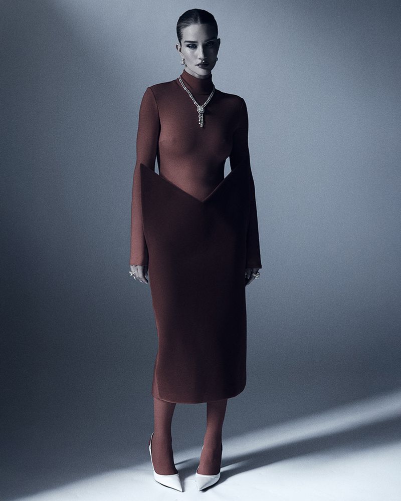 Fashion portrait of a model wearing a long-sleeved, figure-hugging dress with a statement necklace and white pointed heels on a studio backdrop.