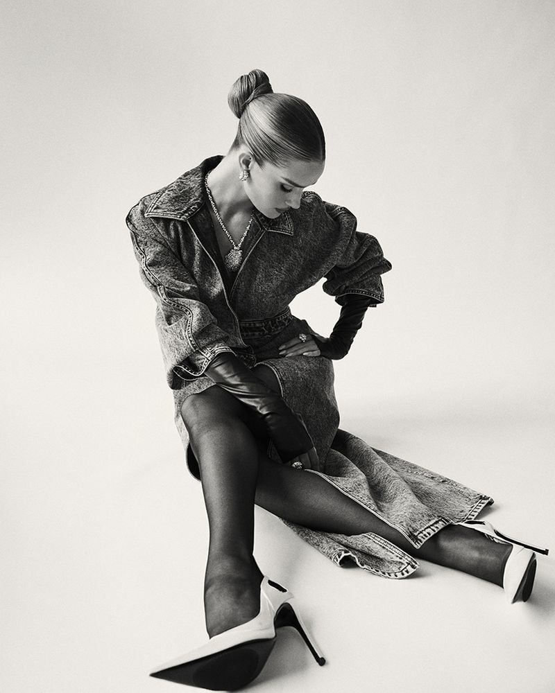 A woman in a denim dress and black tights poses against a white background, showcasing high heels, jewelry, and an elegant updo hairstyle.