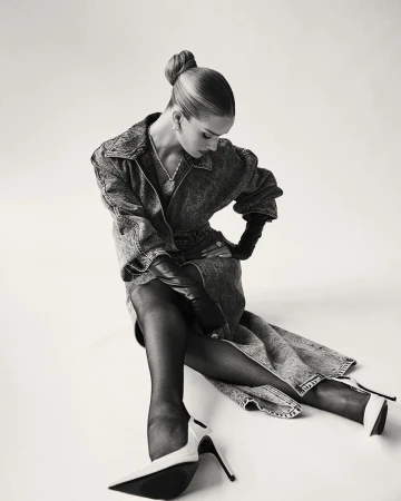 A woman in a denim dress and black tights poses against a white background, showcasing high heels, jewelry, and an elegant updo hairstyle.