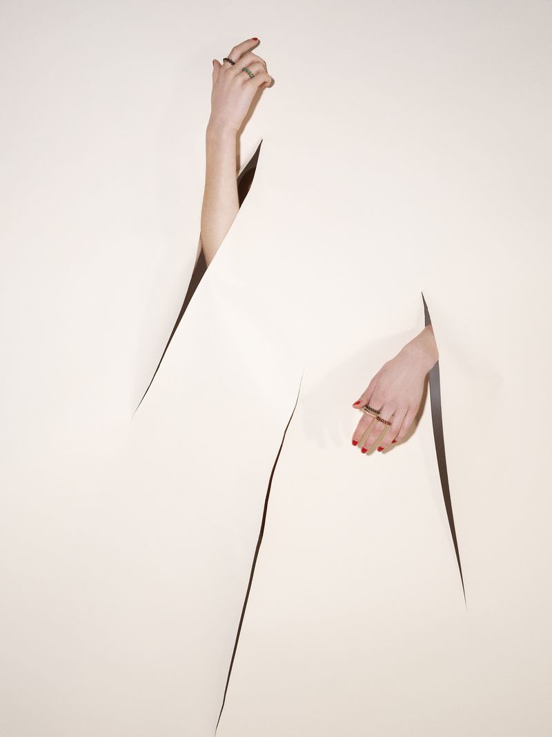 Surreal fashion shot of two hands with red manicured nails and rings poking through geometric cutouts in a light-cream paper surface.