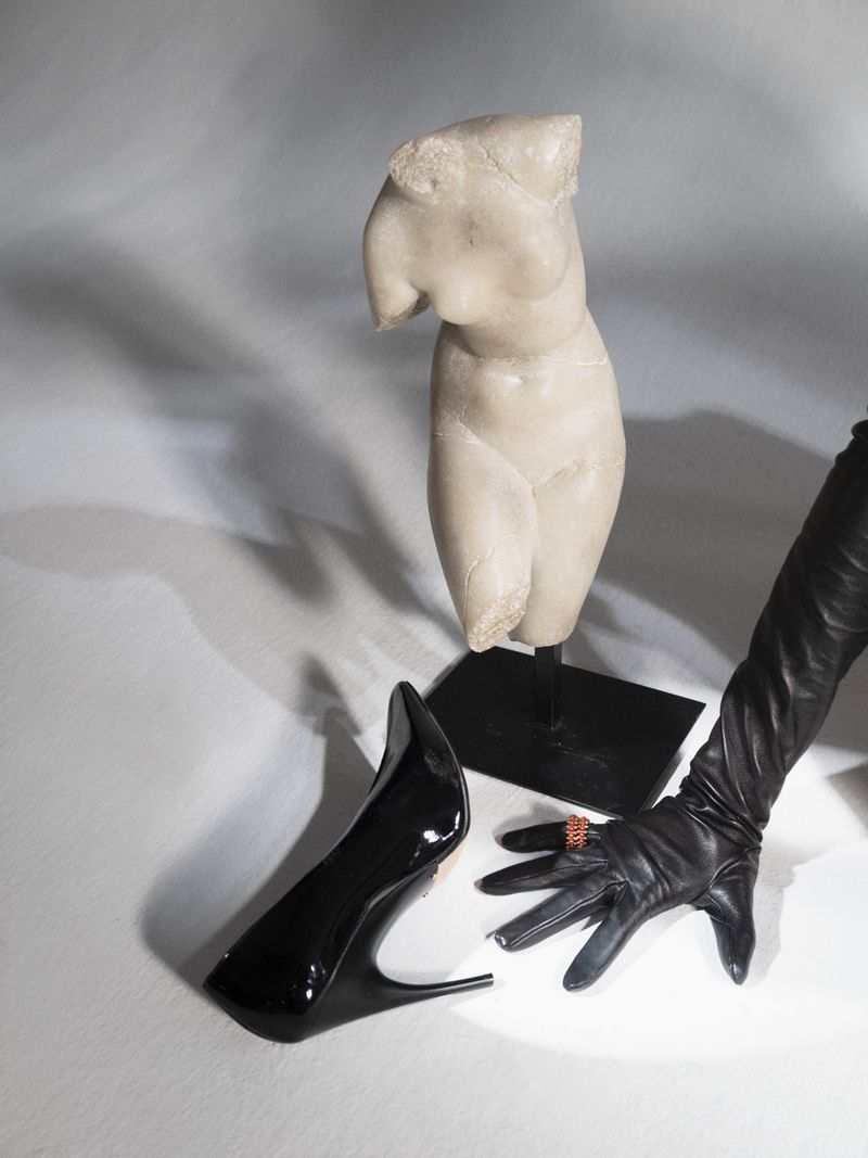 Classical torso sculpture next to a black high heel and long black leather glove accessorized with a gold ring, studio lit.