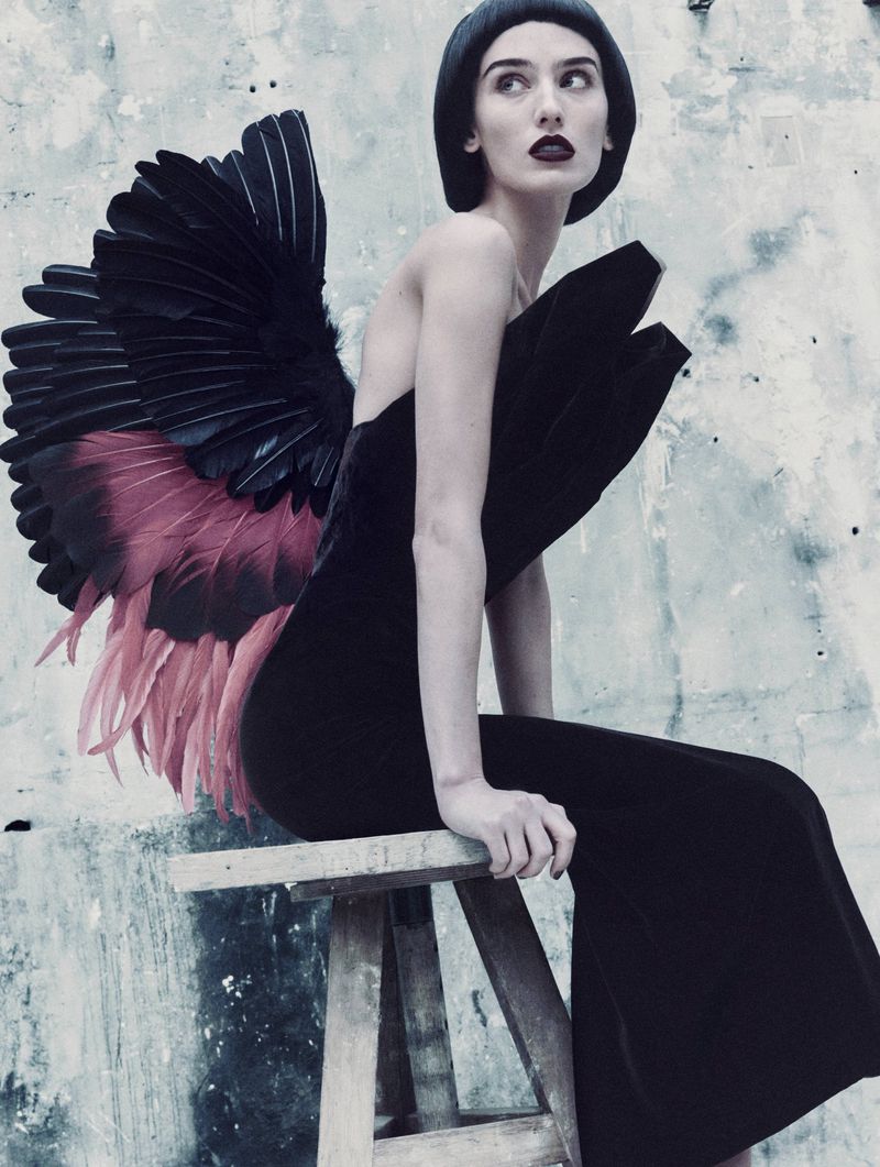 Elegant woman in a black gown and wing-like details seated on a wooden stool, photographed in a moody, textured setting.