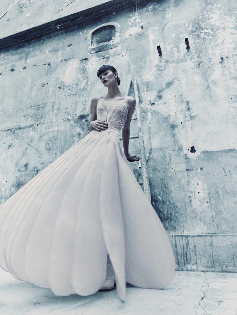 A woman in an elegant white gown poses gracefully against a weathered concrete wall with a ladder. Fashion editorial photography.