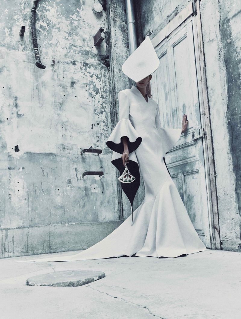 Fashion photography of a woman in a white gown and hat posing in an urban setting with a concrete wall and door in the background.