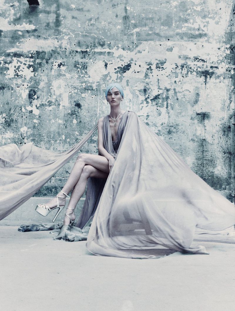 Elegant woman in a long flowing gown and platform heels sits against a weathered wall, showcasing a blend of high fashion and urban decay.
