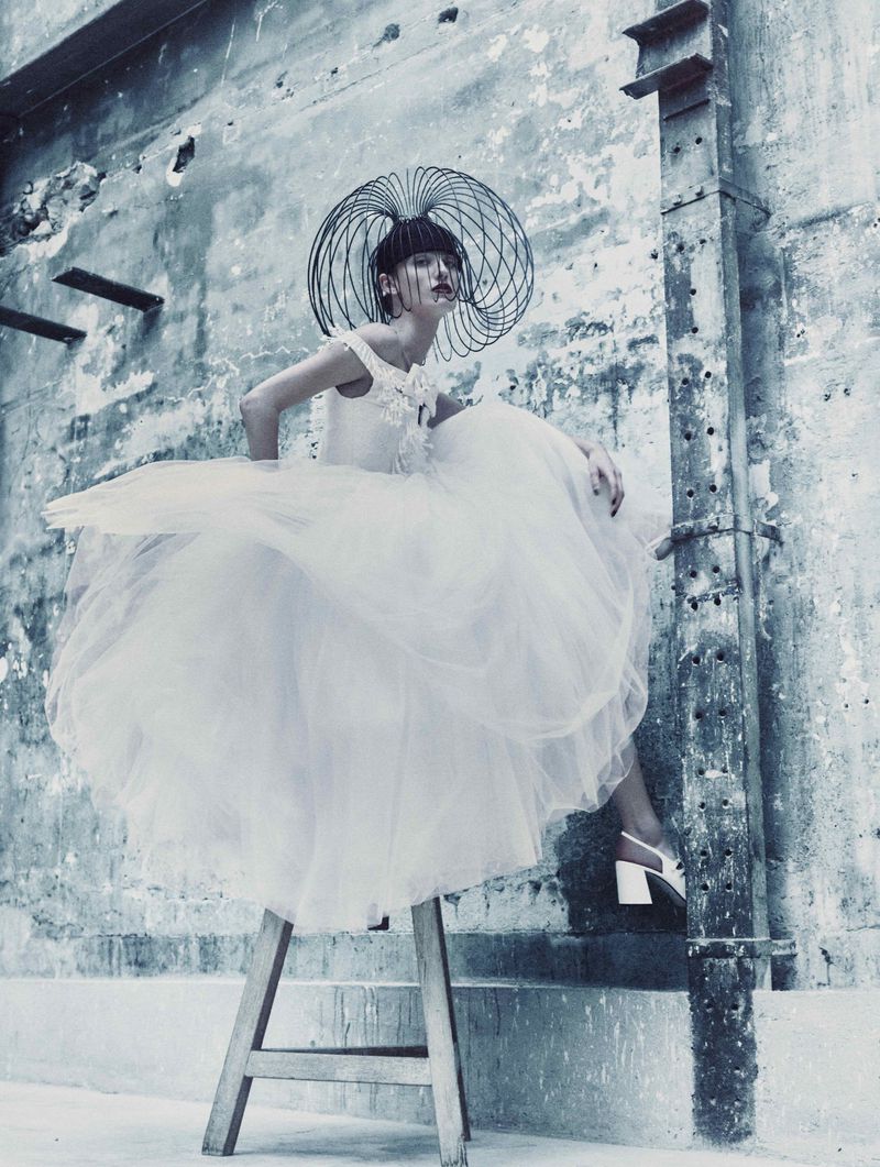 Woman in an off-shoulder dress with a tulle skirt and a sculptural hat sitting on a wooden ladder against a textured wall.