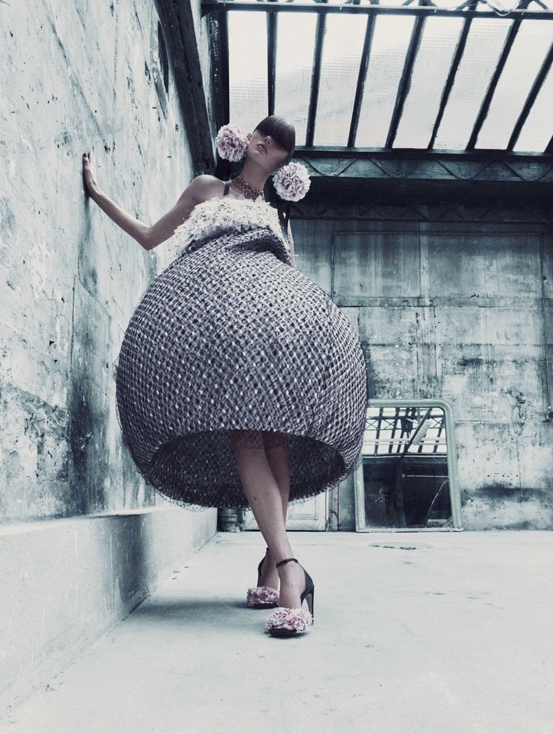 A woman in a large, round dress poses against a concrete wall with flowers in her hair and on her shoes in an industrial setting.