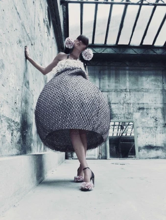 A woman in a large, round dress poses against a concrete wall with flowers in her hair and on her shoes in an industrial setting.