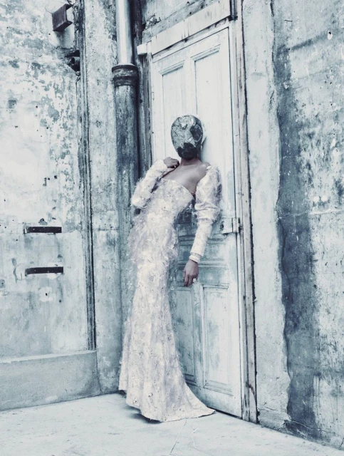 A woman in a white gown stands in front of a weathered door, with a surreal textured covering obscuring her face in a fashion editorial.