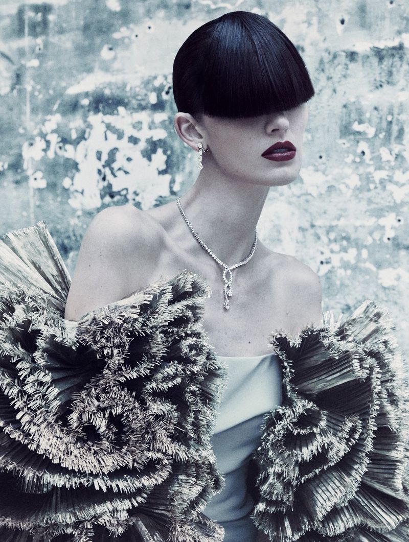 A model poses in a unique ruffled collar with a black bob, dramatic makeup, and jewelry against a mottled blue background.