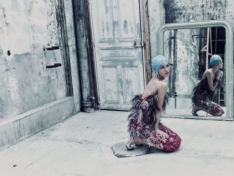 A woman with blue hair and a feathered dress kneels in an urban setting, with a mirror reflecting her image, creating a surreal and stylish scene.