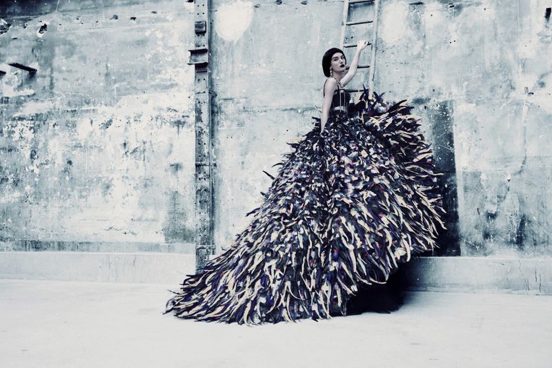 A woman in a feather gown poses against a weathered wall with a ladder, creating an elegant, high-fashion image with a moody aesthetic.